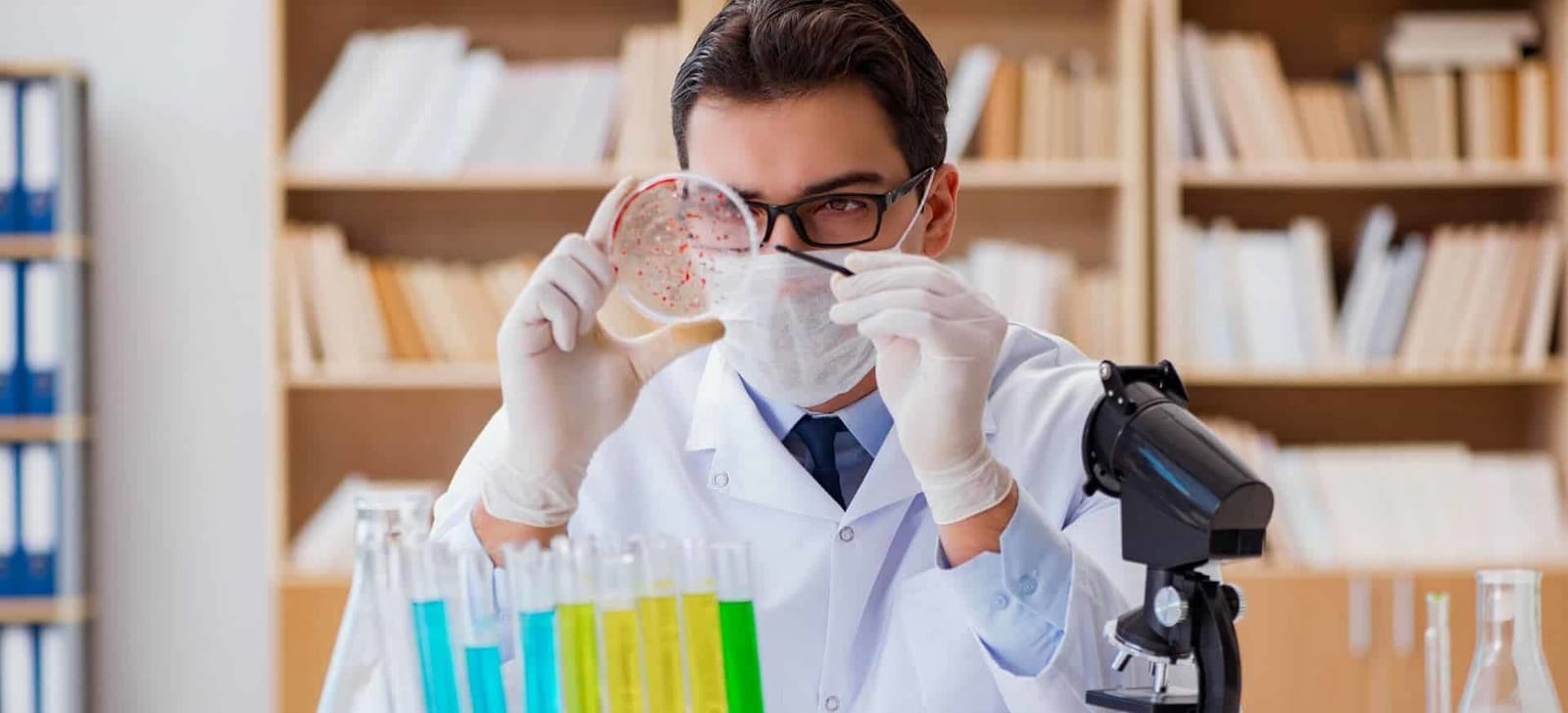Osoba w odzieży laboratoryjnej oglądająca szalkę obok mikroskopu