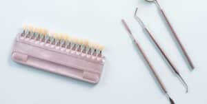 Obraz przedstawia zestaw narzędzi dentystycznych: bloczek z sztucznymi zębami służący do dobierania ich kolorów oraz kilka metalowych narzędzi dentystycznych układających się na białym tle.