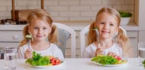 The painting shows two smiling little girls with long blonde hair sitting at a kitchen table. In front of them are plates of vegetable salad. Each of the girls is holding a fork, preparing to eat. The scene appears to depict two sisters or twins at a meal in a domestic kitchen.