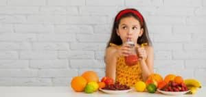 The painting depicts a girl drinking juice, surrounded by various fruits. The girl is wearing a yellow floral dress and a red headband. In front of her are plates of fruit, such as oranges, bananas, grapes and lemons, set against a white background of a brick wall.