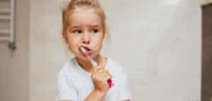 The photograph shows a little girl with fair hair brushing her teeth. The child has her mouth open and is holding a toothbrush to her mouth, performing the act of brushing her teeth. A pink decoration is visible on the girl's white shirt. The interior appears to be a bathroom or children's room.