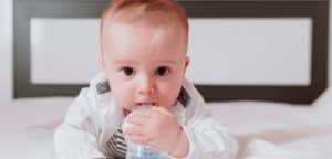 The photograph shows an infant on a bed. The baby has red hair and big brown eyes. He is sucking his little fists, looking directly into the camera lens. He is dressed in a white striped jumpsuit. The background is a white wooden baby bed.
