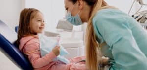 Obraz przedstawia małą dziewczynkę w różowej bluzie siedząca na fotelu dentystycznym oraz dentystkę w niebieskim fartuchu medycznym i masce pochylającą się nad nią i prawdopodobnie pokazującą jej narzędzia dentystyczne. Dziewczynka szeroko się uśmiecha, podczas gdy dentysta sprawdza jej zęby. Scena rozgrywa się w gabinecie stomatologicznym.