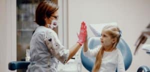 Zdjęcie przedstawia wizytę dziecka u dentysty. Widzimy małą dziewczynkę siedzącą na fotelu stomatologicznym, która przybija piątkę dentystce w okularach ochronnych i masce. Na zdjęciu widoczne są elementy wyposażenia gabinetu stomatologicznego. Scena ta pokazuje pozytywną interakcję między lekarzem a małą pacjentką podczas wizyty kontrolnej.