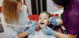 Zdjęcie przedstawia dwie dentystki wykonujące zabieg stomatologiczny na pacjentce, którą jest mała dziewczynka. Dziewczynka siedzi na krześle dentystycznym z otwartymi ustami, podczas gdy dentystki w niebieskich rękawiczkach pracują nad jej uzębieniem używając narzędzi stomatologicznych. Scena rozgrywa się w gabinecie dentystycznym.