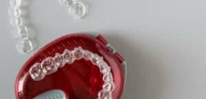 Obraz przedstawia czerwony plastikowy pojemnik używany w gabinetach dentystycznych do przechowywania protez zębowych lub aparatów ortodontycznych. Wewnątrz pojemnika znajduje się przezroczysta, gumowa dziąsłowa półłukowa podpórka do stabilizacji protez. Obok pojemnika leżą przezroczyste elastyczne rozpórki, prawdopodobnie służące do rozszerzania szczęk w procesie leczenia ortodontycznego.