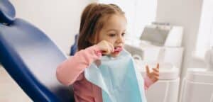 Obraz przedstawia małą dziewczynkę siedzącą na dentystycznym fotelu w jasnym gabinecie stomatologicznym. Dziecko ma rozpuszczone rude włosy i ma na sobie różową bluzeczkę. Wydaje się zainteresowane sytuacją i spogląda w kierunku aparatury dentystycznej widocznej na drugim planie. Scena nawiązuje do rutynowej wizyty u dentysty przez małą pacjentkę.