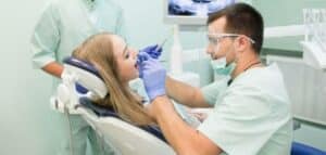 The image shows a dentist in full surgical protective gear, including mask, goggles and gloves, performing a dental procedure on a young patient lying in a dental chair. We also see the interior of a professional dental office with medical equipment.