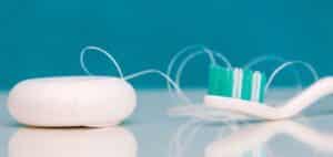 Obraz przedstawia szczoteczkę do zębów i nitkę dentystyczną na błękitnym, lekko lśniącym tle. Szczoteczka ma zielono-białe włosie, a nitka dentystyczna jest podłużnego, walcowatego kształtu w kolorze białym. Przedmioty leżą na gładkiej powierzchni, która odbija ich kształty. Obraz jest skoncentrowany na przyborach higienicznych wykorzystywanych do pielęgnacji jamy ustnej.