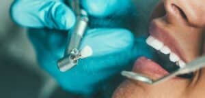 Zdjęcie przedstawia wizytę u dentysty. Widoczne są rękawiczki w kolorze niebieskim, narzędzia stomatologiczne oraz szerokie uśmiechnięte usta pacjenta z białymi zębami. Obraz ilustruje typową scenę leczenia stomatologicznego z bliska.