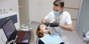 Imaginea prezintă un dentist care examinează un pacient într-un cabinet stomatologic. Dentistul, purtând un șorț alb și o mască, se uită atent la gura deschisă a pacientului așezat pe scaun. Masa prezintă instrumente dentare și un monitor de computer.