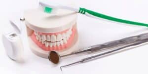 Zdjęcie przedstawia przybory dentystyczne: model uzębienia, szczoteczkę do zębów, lustrzadełko stomatologiczne oraz sondę. Ujęcie skupia się na przyrządach wykorzystywanych przez dentystów podczas leczenia pacjentów.