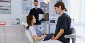 Obraz przedstawia gabinet dentystyczny. Widać pacjentkę siedzącą na fotelu stomatologicznym, dentyskę stojącą obok niej oraz uśmiechniętego dentystę w tle. Pacjentka i dentyści są uśmiechnięci, a atmosfera wydaje się przyjazna i profesjonalna.