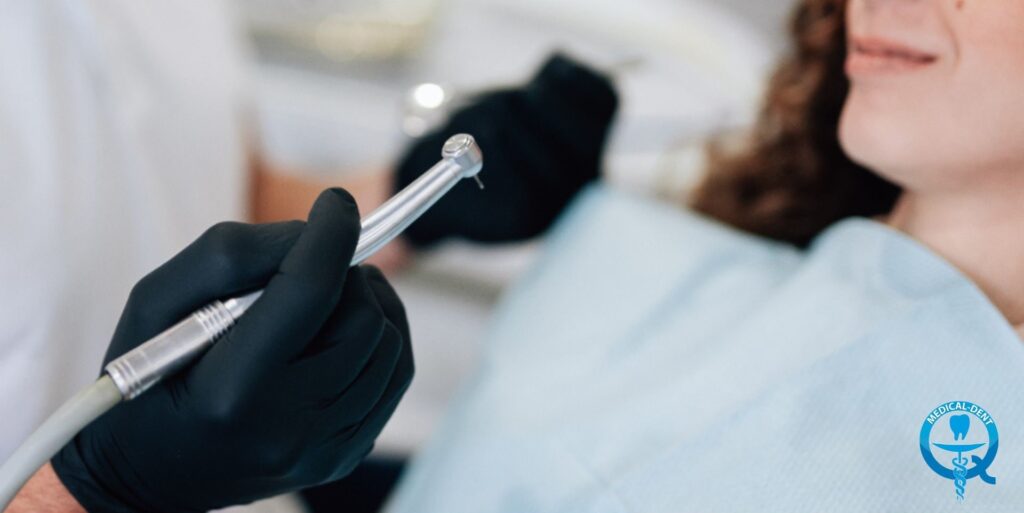 The photograph shows the dentist's hand holding a dental tool, probably a drill, ready to work on the patient's teeth. We can see half of the patient's face and the medical logo on the apron. The image has a shallow depth of field, focusing on the dental tool.
