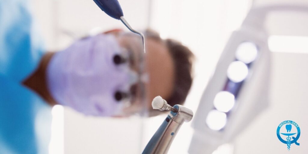 The image shows a child undergoing dental treatment. A young child can be seen lying on a dentist's chair while the dentist uses a dental instrument to work in his mouth. In the background are blurred, fuzzy lights, suggesting that the scene is taking place in a dentist's office.
