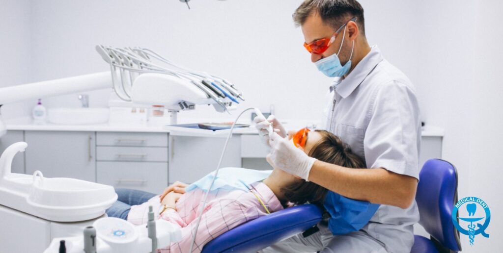 Dentysta w białym fartuchu, w masce i pomarańczowych okularach ochronnych, przeprowadza zabieg na pacjencie leżącym na niebieskim fotelu dentystycznym. W tle widoczny jest standardowy sprzęt stomatologiczny, w tym unit dentystyczny z końcówkami oraz szafki medyczne w jasnym gabinecie.