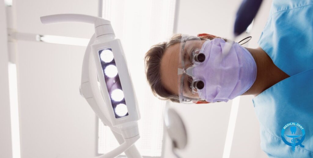 Dentysta w masce ochronnej i okularach powiększających podczas zabiegu. W kadrze widoczna profesjonalna lampa dentystyczna LED w kolorze białym. W tle jasne, sterylne pomieszczenie gabinetu stomatologicznego.