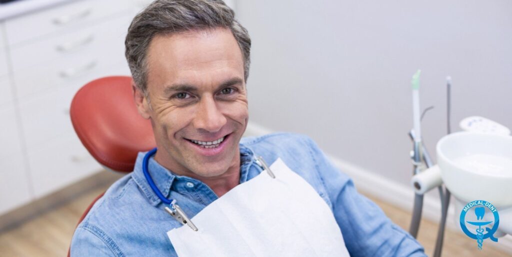"Un pacient zâmbitor într-o cămașă din denim stă într-un scaun dentar roșu. În fundal, echipamentul dentar și interiorul luminos și steril al cabinetului sunt vizibile. În colțul din dreapta jos se află logo-ul medical în albastru."