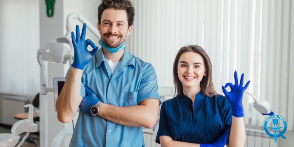 Personalul stomatologic dintr-un cabinet stomatologic, purtând îmbrăcăminte medicală profesională și mănuși albastre, arată gestul "OK". În fundal, sunt vizibile echipamentele dentare și interiorul luminos al cabinetului. În colțul din dreapta jos se află logo-ul medical.