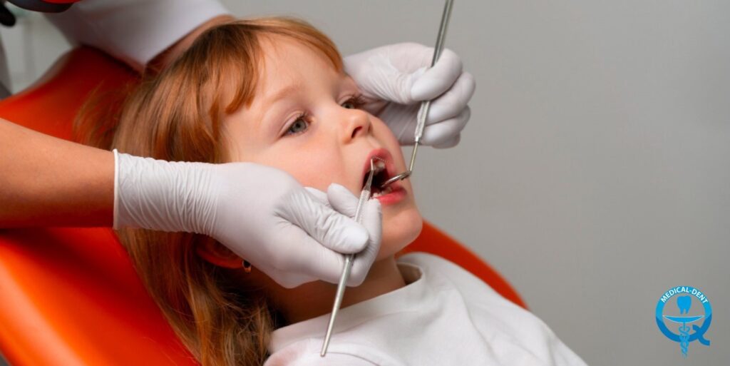 Małe dziecko podczas badania stomatologicznego, leżące na pomarańczowym fotelu dentystycznym. Dentysta w białych rękawiczkach używa narzędzia dentystycznego do badania zębów. W prawym dolnym rogu widoczne jest niebieskie logo medyczne.