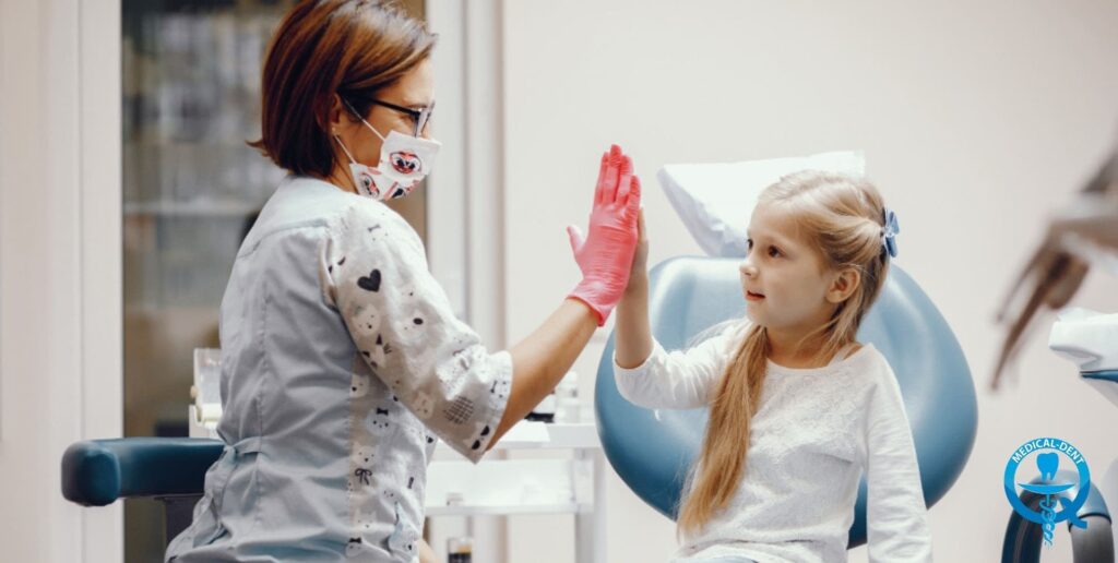 Dentysta w ozdobnym fartuchu i maseczce przybija piątkę z małą pacjentką siedzącą na fotelu dentystycznym. Dziewczynka ma długie jasne włosy z kokardką i uśmiecha się. Gabinet jest jasny i nowocześnie wyposażony.