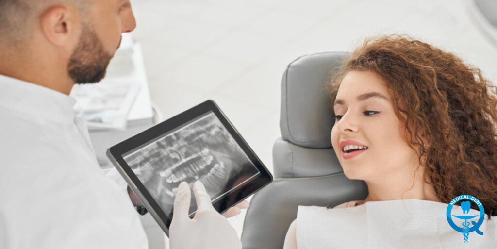 Pacjentka z kręconymi włosami siedzi w fotelu dentystycznym, podczas gdy dentysta w białym fartuchu pokazuje jej zdjęcie rentgenowskie zębów na tablecie. W prawym dolnym rogu widoczne logo gabinetu stomatologicznego.