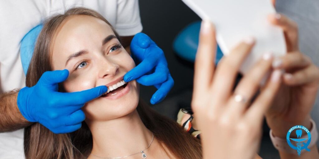 Pacjentka podczas badania stomatologicznego, uśmiechająca się i przeglądająca swoje zęby w lusterku. Dentysta w niebieskich rękawiczkach delikatnie odsuwa jej policzek. W prawym dolnym rogu widoczne logo kliniki stomatologicznej Medical-Dent.