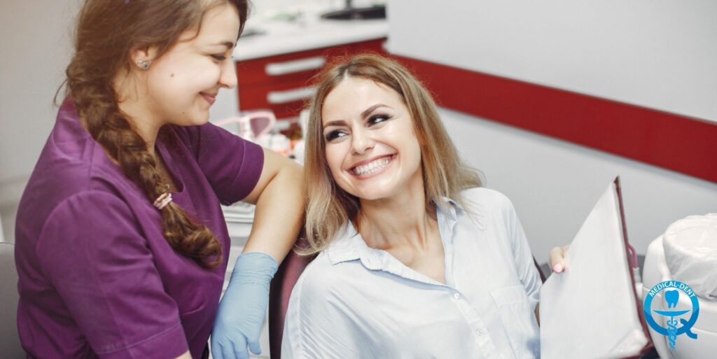 Smiling patient in dental surgery accompanied by dentist in purple uniform with blue gloves. Red dental furniture visible in the background. In the lower right corner the logo of the dental clinic.