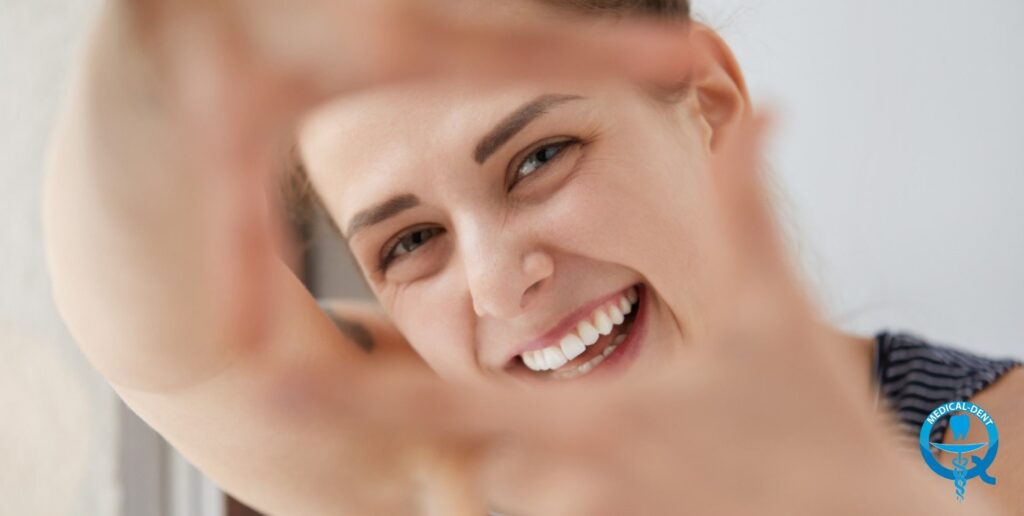 "A person smiling, showing healthy white teeth, viewed through a frame formed by a hand. The logo of the Medical-Dent dental clinic is visible in the lower right corner."