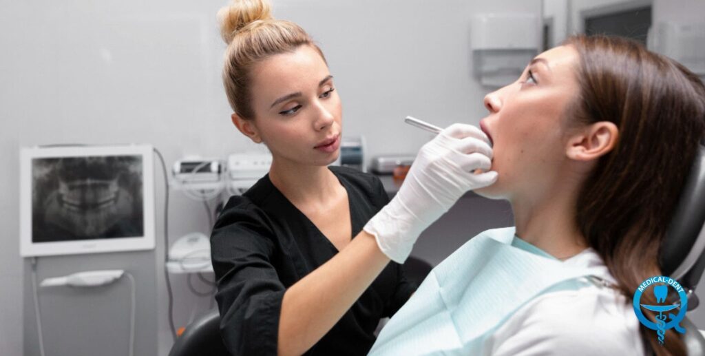 A dentist wearing white protective gloves examines the teeth of a patient lying in a dental chair in a modern dental office with medical equipment visible in the background.