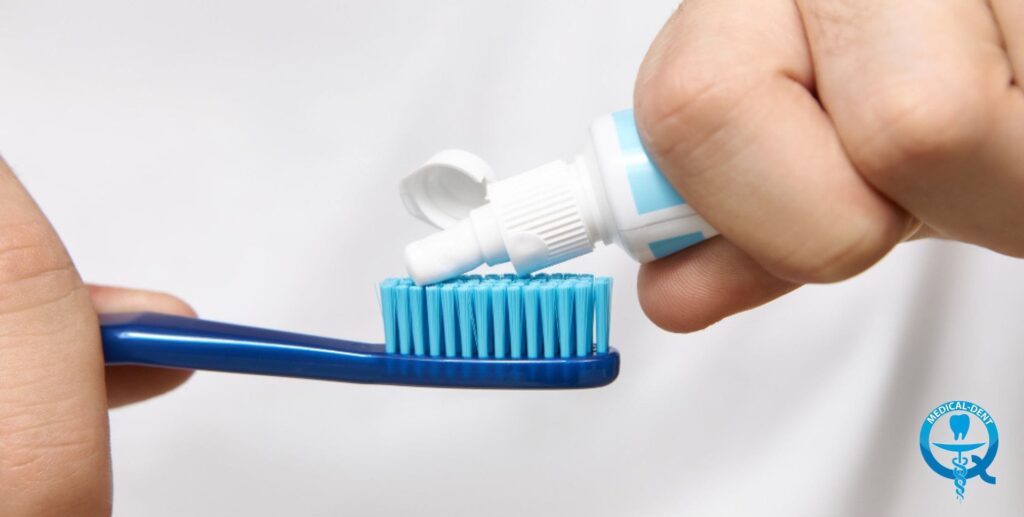 Dłonie wyciskające pastę do zębów z tuby na niebieską szczoteczkę do zębów, na białym tle, z logo Medical-Dent w rogu.