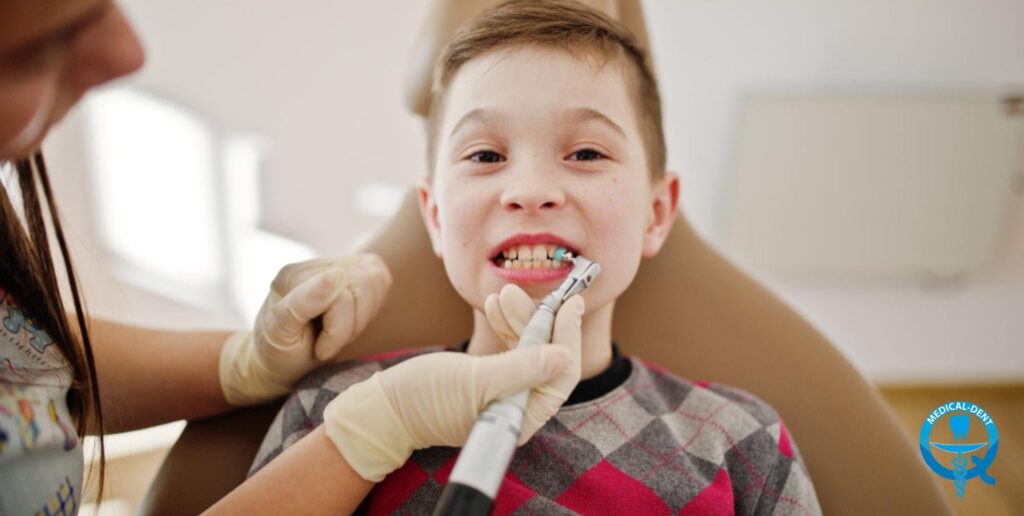 Chłopiec siedzący na fotelu dentystycznym podczas czyszczenia zębów przez dentystę w rękawiczkach używającego polerki stomatologicznej, uśmiechający się do aparatu.