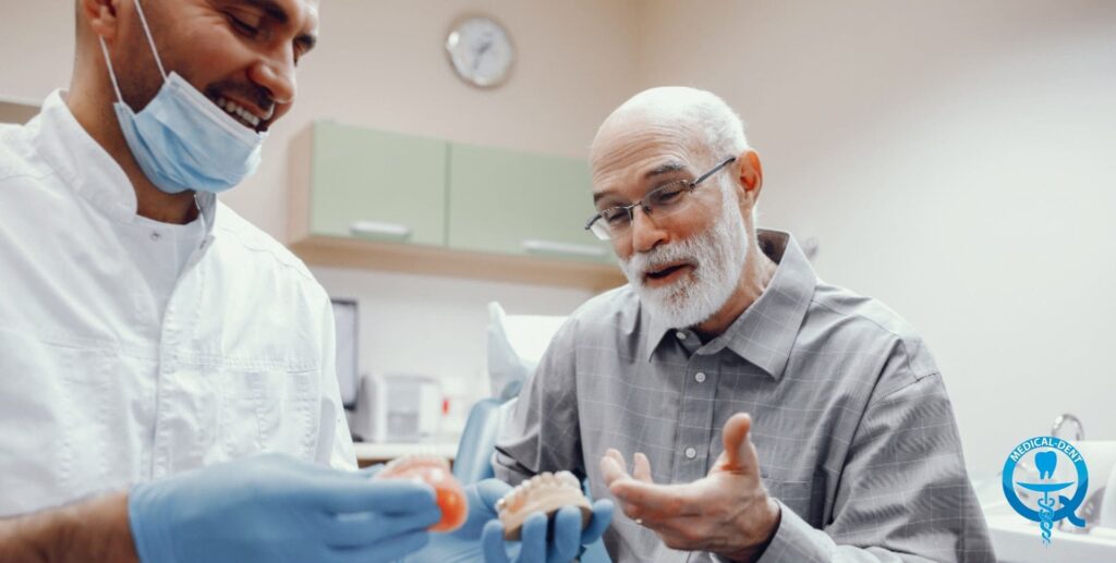 Dentysta w białym fartuchu i masce ochronnej pokazuje starszemu pacjentowi w okularach model szczęki z protezą zębową w gabinecie stomatologicznym w Wielkiej Brytanii.