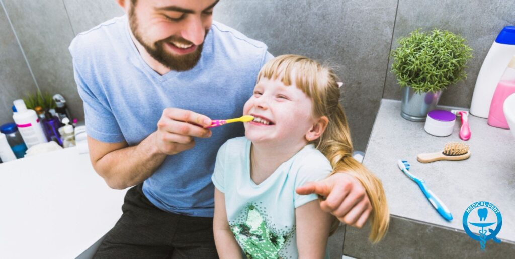 Усмихнат баща мие зъбите на щастливо момиченце в банята, държейки жълта четка за зъби. На заден план се виждат тоалетни принадлежности и логото на Medical-Dent.