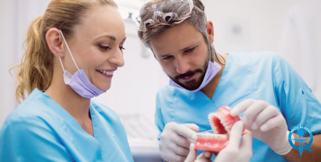 Doi medici stomatologi în șorțuri albastre și măști de protecție mov se uită la o proteză dentară temporară în cabinetul unui dentist. Bărbatul, care poartă mănuși, ține proteza roz, în timp ce femeia privește munca cu zâmbetul pe buze.