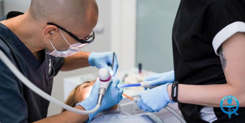 Dentysta w okularach i masce ochronnej, ubrany w niebieskie rękawiczki, wykonuje zabieg stomatologiczny na pacjentce leżącej na fotelu. Asystentka w czarnym fartuchu pomaga, trzymając narzędzia dentystyczne. Gabinet stomatologiczny Medical-Dent w tle.