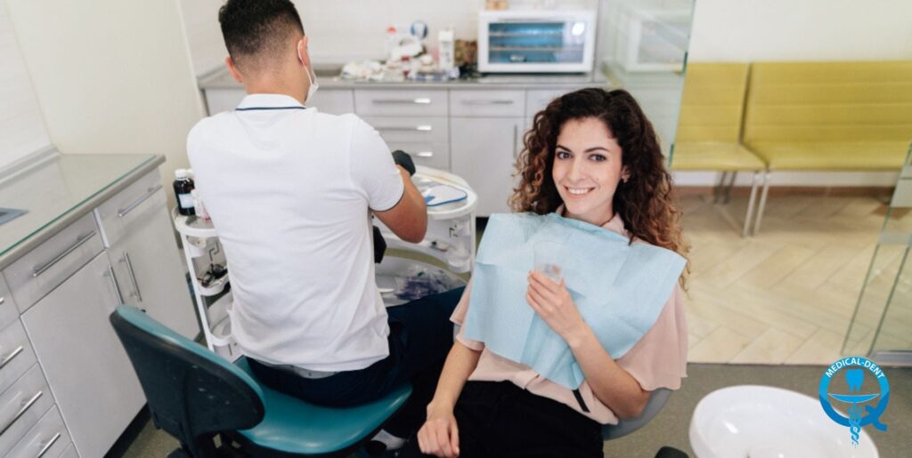 Uśmiechnięta pacjentka z kręconymi włosami siedzi na fotelu dentystycznym z niebieskim śliniaczkiem, podczas gdy dentysta w białym fartuchu i masce przygotowuje narzędzia w nowoczesnym gabinecie stomatologicznym w Wielkiej Brytanii.
