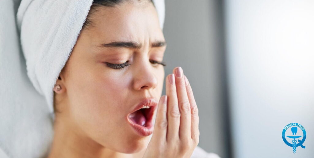 Жена с увита около главата си кърпа проверява дъха си, издишвайки в ръката си с ясно изразена загриженост на лицето - илюстрация на проблема с лошия дъх (халитоза).