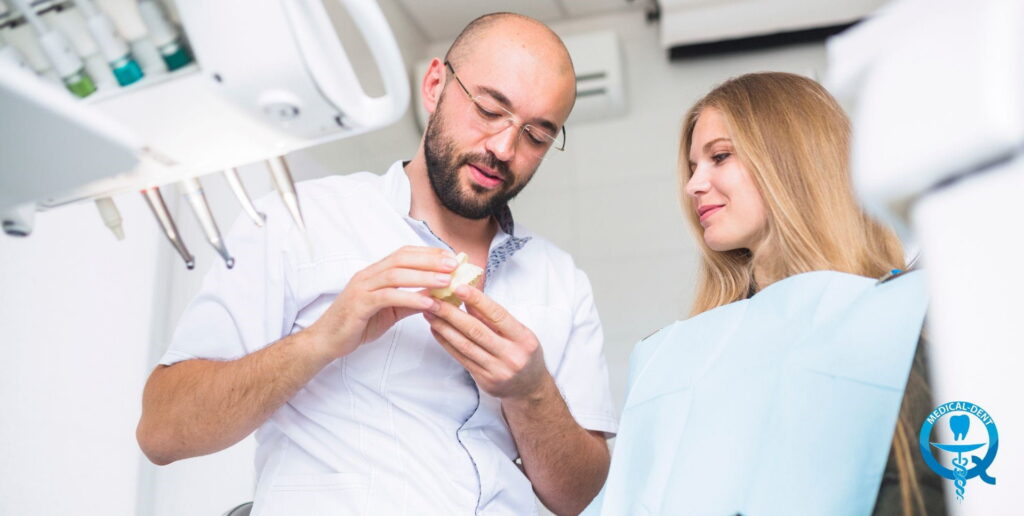 A dentist in a white coat and glasses is showing a patient a model of teeth in a dentist’s surgery; dental instruments and a dentist’s chair can be seen in the background.
