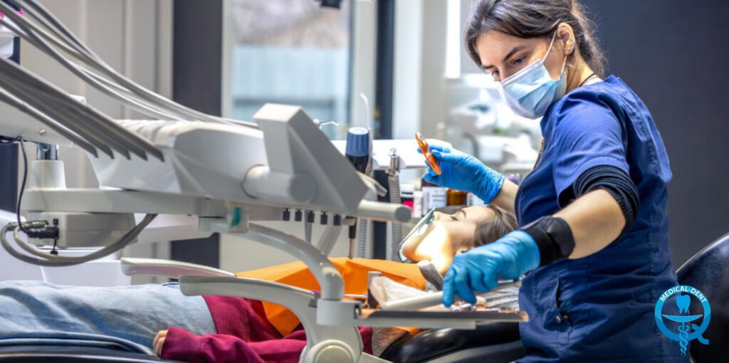 A dentist wearing a blue lab coat and a face shield, with blue gloves, performing a dental procedure on a patient lying in a dental chair in a modern dental surgery in the UK.