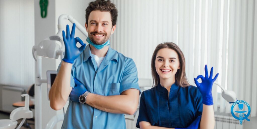 Dvaja usmievaví zubári v modrých uniformách a modrých latexových rukaviciach, ktorí ukazujú gesto „OK", stoja v zubnej ordinácii vo Veľkej Británii.