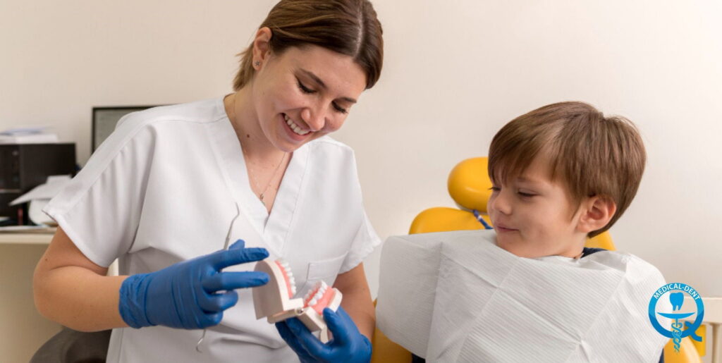 Uśmiechnięta dentystka w białym fartuchu i niebieskich rękawiczkach pokazuje chłopcu model szczęki podczas wizyty stomatologicznej. Dziecko siedzi na żółtym fotelu dentystycznym z białym śliniaczkiem, z zainteresowaniem przyglądając się modelowi zębów.
