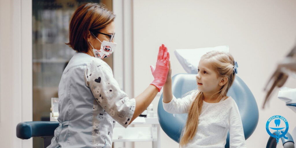 Dentystka w maseczce i różowych rękawiczkach przybija piątkę z małą dziewczynką siedzącą na fotelu dentystycznym w gabinecie stomatologicznym.