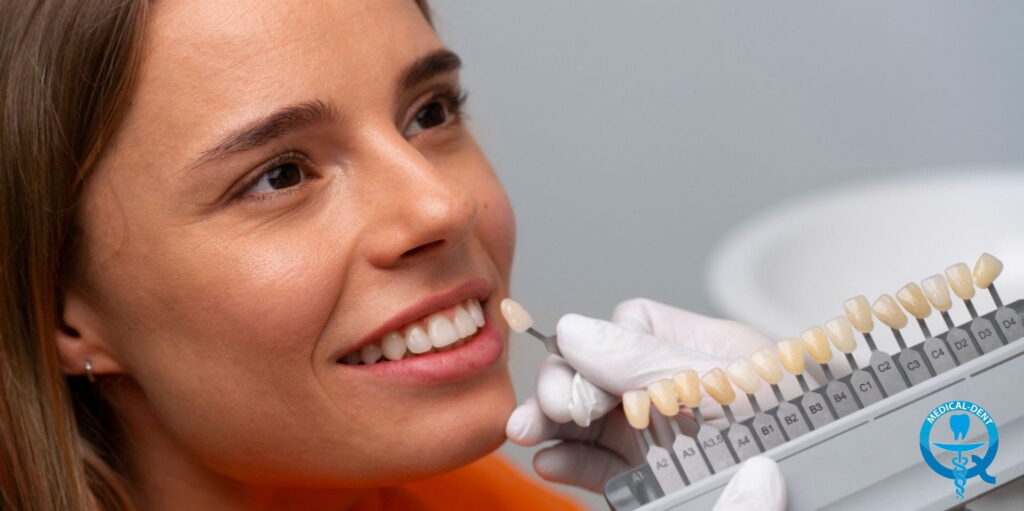 Uśmiechnięta kobieta na fotelu dentystycznym podczas dobierania koloru zębów – dentysta w rękawiczkach przykłada próbnik odcieni do jej zębów, trzymając skalę kolorów porcelanowych koron dentystycznych.