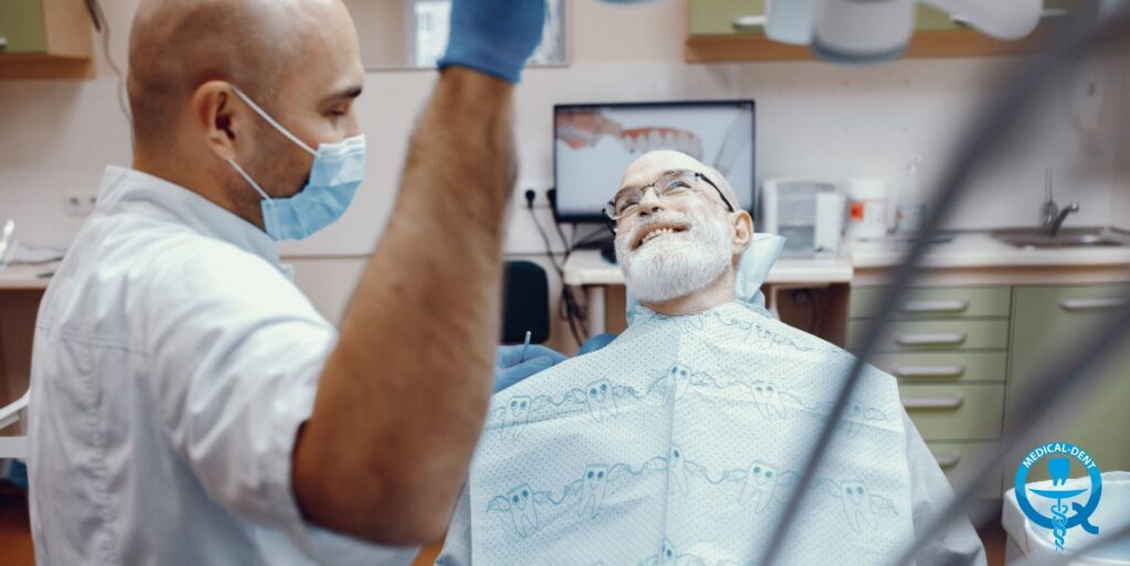 Dentysta w masce chirurgicznej i niebieskich rękawiczkach przeprowadza zabieg stomatologiczny u uśmiechniętego starszego mężczyzny z siwą brodą i okularami, siedzącego na fotelu dentystycznym z założonym śliniaczkiem, w nowoczesnym gabinecie stomatologicznym.