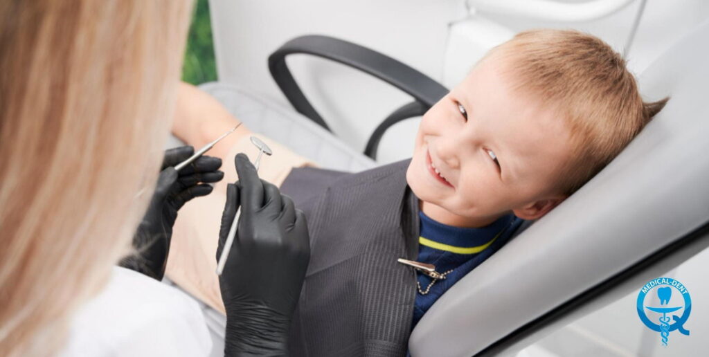Uśmiechnięty chłopiec siedzący na fotelu dentystycznym podczas wizyty u dentysty w Wielkiej Brytanii, z dentystką w czarnych rękawiczkach trzymającą narzędzia stomatologiczne.